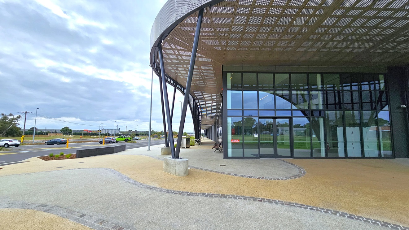 A concrete pavement adorns the concourse, with vehicle parking on the far left. In the middle stands a modern facility building of concrete and glass, featuring a large circular shade with steel mesh. The entrance boasts a floor-to-ceiling glass wall with an automatic door. Chairs and bike racks are visible on the left side. A concrete pavement adorns the concourse, with vehicle parking on the far left. In the middle stands a modern facility building of concrete and glass, featuring a large circular shade with steel mesh. The entrance boasts a floor-to-ceiling glass wall with an automatic door. Chairs and bike racks are visible on the left side.