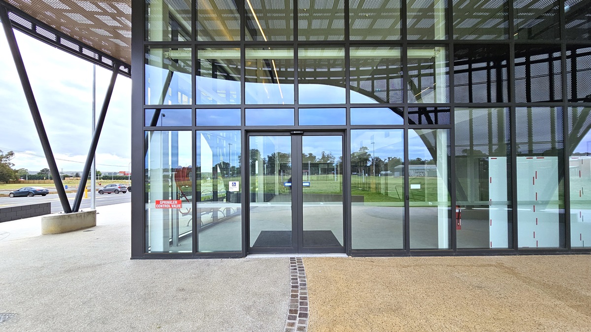 A concrete pavement adorns the concourse, with vehicle parking on the far left. In the middle stands a modern facility building of concrete and glass, featuring a large circular shade with steel mesh. The entrance boasts a floor-to-ceiling glass wall with an automatic sliding door. Chairs and bike racks are visible on the left side. A concrete pavement adorns the concourse, with vehicle parking on the far left. In the middle stands a modern facility building of concrete and glass, featuring a large circular shade with steel mesh. The entrance boasts a floor-to-ceiling glass wall with an automatic sliding door. Chairs and bike racks are visible on the left side.
