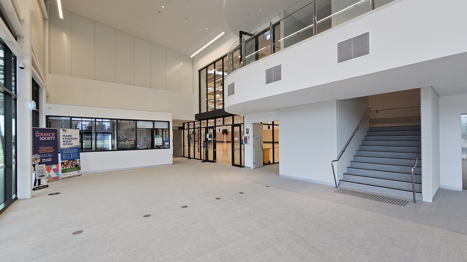 To the right is a staircase with handrails and truncated domes. Inside, a high ceiling accommodates a mezzanine. Straight ahead is the reception desk. Retractable banners are positioned on the left, near the glass window. To the right of the reception is the stadium, secured with glass walls. The floor boasts a firm vinyl finish. To the right is a staircase with handrails and truncated domes. Inside, a high ceiling accommodates a mezzanine. Straight ahead is the reception desk. Retractable banners are positioned on the left, near the glass window. To the right of the reception is the stadium, secured with glass walls. The floor boasts a firm vinyl finish.