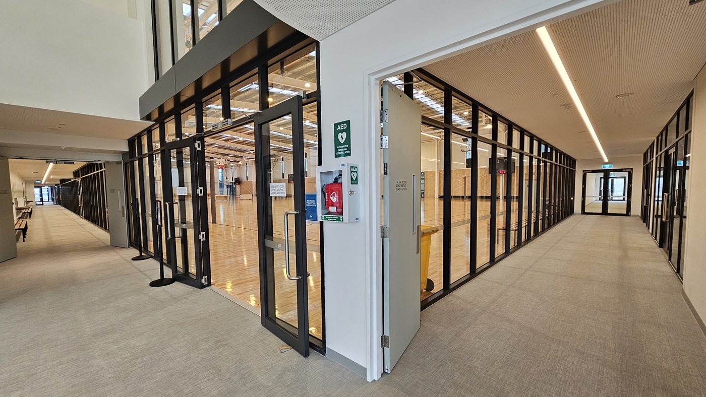The stadium entrance features a manual outward-opening glass door with wall signage, including a defibrillator. To the right, a corridor leads to toilets, lockers, and change rooms. On the left, another corridor leads to bathrooms, Change Places, and the Function Room. Bench chairs and drinking fountains are mounted on the wall. The wall is adorned with foliage decoration, while the ceiling is white. The floor is firm vinyl. The stadium entrance features a manual outward-opening glass door with wall signage, including a defibrillator. To the right, a corridor leads to toilets, lockers, and change rooms. On the left, another corridor leads to bathrooms, Change Places, and the Function Room. Bench chairs and drinking fountains are mounted on the wall. The wall is adorned with foliage decoration, while the ceiling is white. The floor is firm vinyl.
