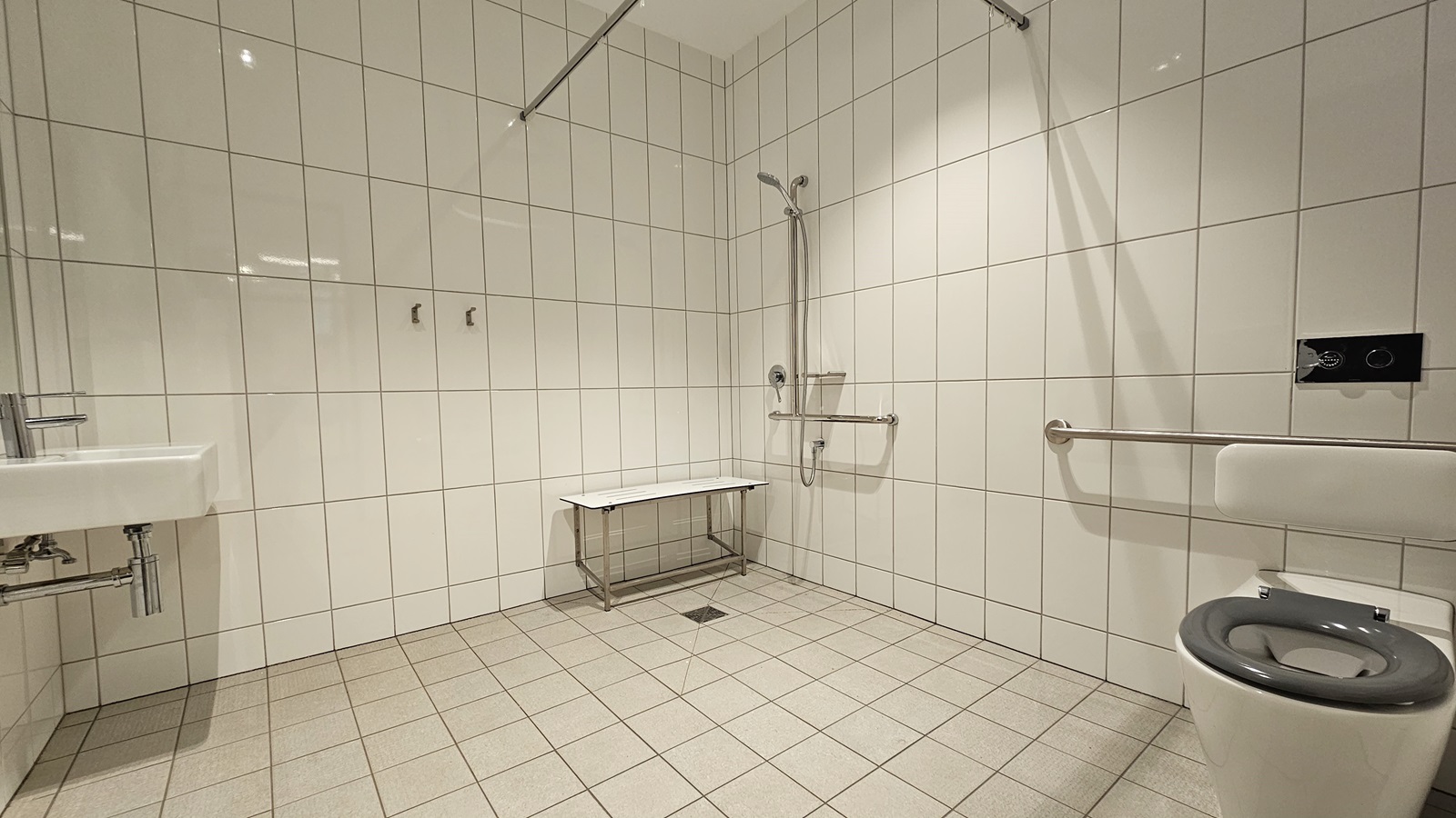 Inside the accessible toilet, a wash basin is positioned to the left. Next to it is a shower with hand grabs and a foldable bench chair. To the right is a toilet bowl with hand grabs. White tiles cover the entire floor and wall. Above, a placeholder for a curtain hangs in place. Inside the accessible toilet, a wash basin is positioned to the left. Next to it is a shower with hand grabs and a foldable bench chair. To the right is a toilet bowl with hand grabs. White tiles cover the entire floor and wall. Above, a placeholder for a curtain hangs in place.