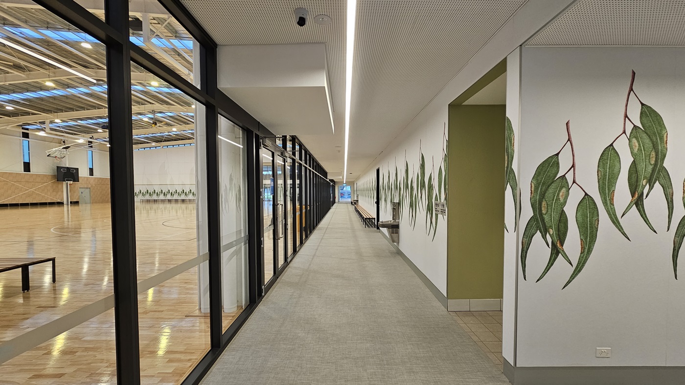 To the left is the stadium enclosed by glass, revealing polished timber floors and an exposed industrial ceiling. In the middle is a corridor leading to the reception area. To the right is a white wall with green foliage decorations extending into the stadium. Adjacent is a tiled area leading to an accessible toilet, marked by signage. The ceiling is white with installed CCTV, and the floor has firm vinyl. To the left is the stadium enclosed by glass, revealing polished timber floors and an exposed industrial ceiling. In the middle is a corridor leading to the reception area. To the right is a white wall with green foliage decorations extending into the stadium. Adjacent is a tiled area leading to an accessible toilet, marked by signage. The ceiling is white with installed CCTV, and the floor has firm vinyl.