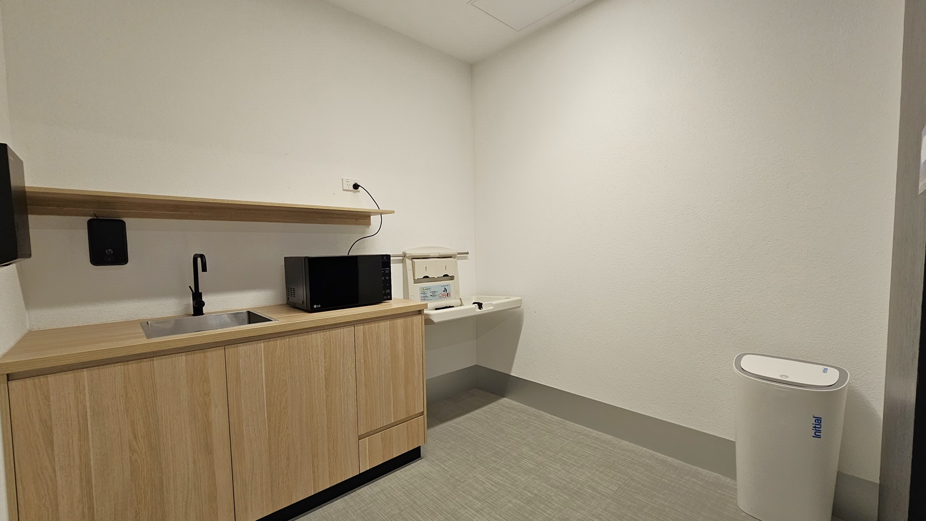 On the left, there's a wash basin with a wooden cabinet and ledge. A microwave oven sits on top. Soap and tissue dispensers are fixed to the wall. A white rubbish bin stands on the right. The ceiling is white, and the floor is firm vinyl. On the left, there's a wash basin with a wooden cabinet and ledge. A microwave oven sits on top. Soap and tissue dispensers are fixed to the wall. A white rubbish bin stands on the right. The ceiling is white, and the floor is firm vinyl.