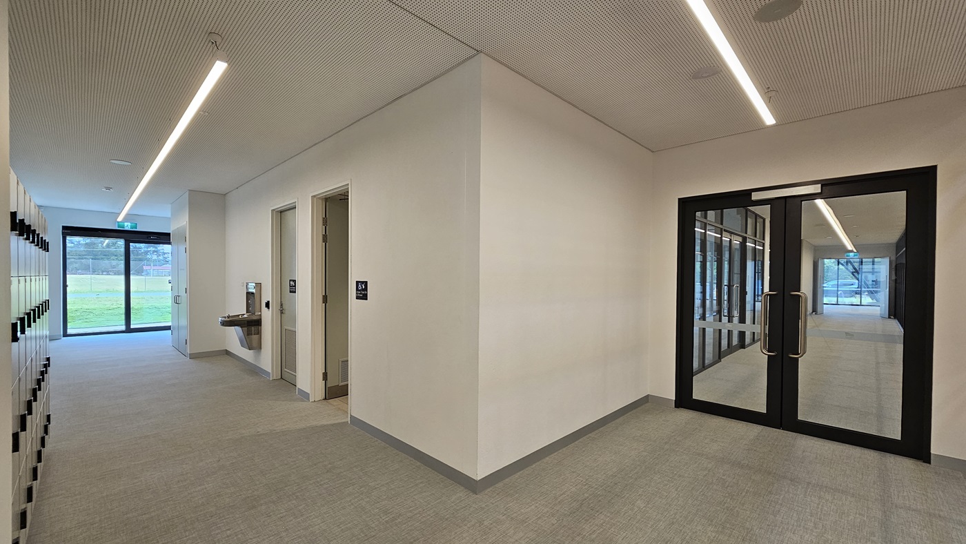 On the left, there's a 4-tier locker area. In the middle, an accessible toilet is marked with a gender-access sign. A drinking faucet is fixed to the wall. At the far end, a glass wall offers an outside view. To the right, a manual glass door. The wall and ceiling are white with LED lights. The floor has firm vinyl. On the left, there's a 4-tier locker area. In the middle, an accessible toilet is marked with a gender-access sign. A drinking faucet is fixed to the wall. At the far end, a glass wall offers an outside view. To the right, a manual glass door. The wall and ceiling are white with LED lights. The floor has firm vinyl.