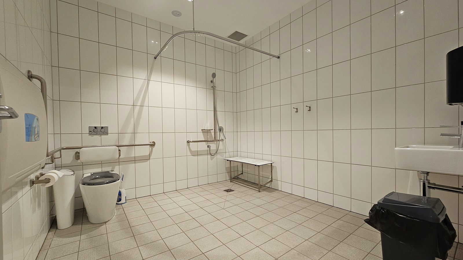 Inside the accessible toilet, a wash basin is on the right with a black rubbish bin nearby. In the middle, a toilet bowl with hand grabs and a shower with a foldable bench chair underneath. On the left, a foldable change table is mounted to the wall next to a rubbish bin and tissue dispenser. White tiles cover the floor and wall, with a curtain holder above. Inside the accessible toilet, a wash basin is on the right with a black rubbish bin nearby. In the middle, a toilet bowl with hand grabs and a shower with a foldable bench chair underneath. On the left, a foldable change table is mounted to the wall next to a rubbish bin and tissue dispenser. White tiles cover the floor and wall, with a curtain holder above.