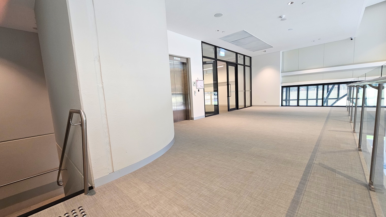 The mezzanine floor includes a lift on the right and stairs with handrails and truncated domes. A view of the lobby below is visible, along with retractable banners in front of the reception. Far ahead is a glass door that opens outward. The floor has vinyl tiles, and the wall and ceiling are all white with a porous design and LED lights. The mezzanine floor includes a lift on the right and stairs with handrails and truncated domes. A view of the lobby below is visible, along with retractable banners in front of the reception. Far ahead is a glass door that opens outward. The floor has vinyl tiles, and the wall and ceiling are all white with a porous design and LED lights.