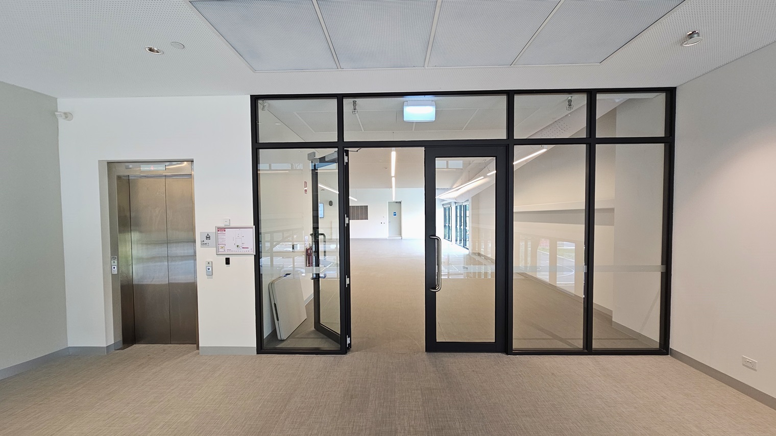 To the left is a lift. Next to it is a manual inward-opening glass door leading to the Function Room, surrounded by glass. On the right is another lift and a manual glass door. A site map is affixed to the wall. A foldable table rests against the left wall. The floor has firm vinyl, and the walls and ceiling are all white. To the left is a lift. Next to it is a manual inward-opening glass door leading to the Function Room, surrounded by glass. On the right is another lift and a manual glass door. A site map is affixed to the wall. A foldable table rests against the left wall. The floor has firm vinyl, and the walls and ceiling are all white.