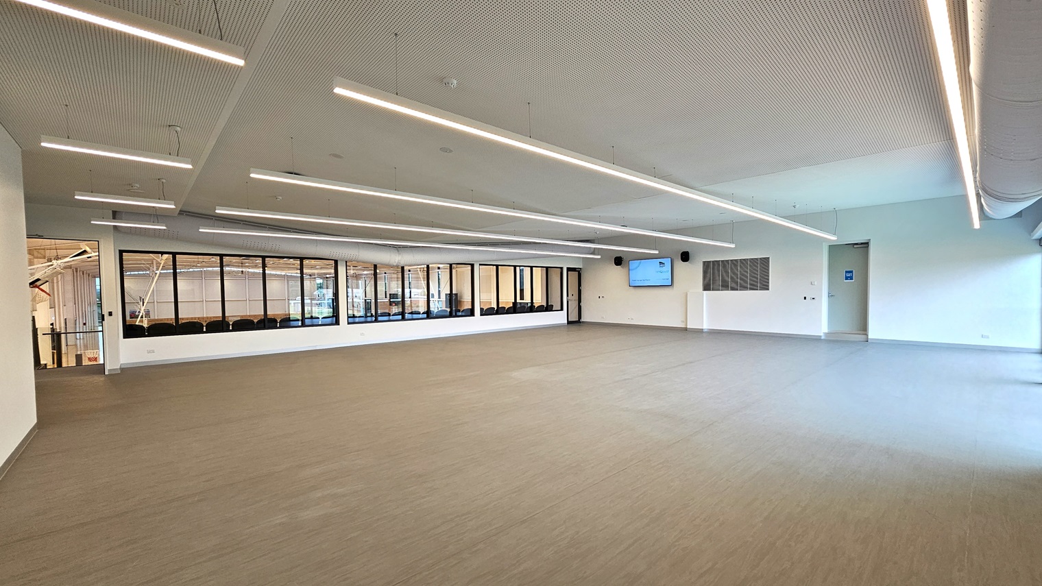 The Function Room is a spacious area with vinyl flooring. A large flatscreen TV is mounted on one wall. Three doors provide access, including one leading to the stadium's spectator area. The walls are white, and the ceiling features a white porous design with fluorescent lighting. The Function Room is a spacious area with vinyl flooring. A large flatscreen TV is mounted on one wall. Three doors provide access, including one leading to the stadium's spectator area. The walls are white, and the ceiling features a white porous design with fluorescent lighting.