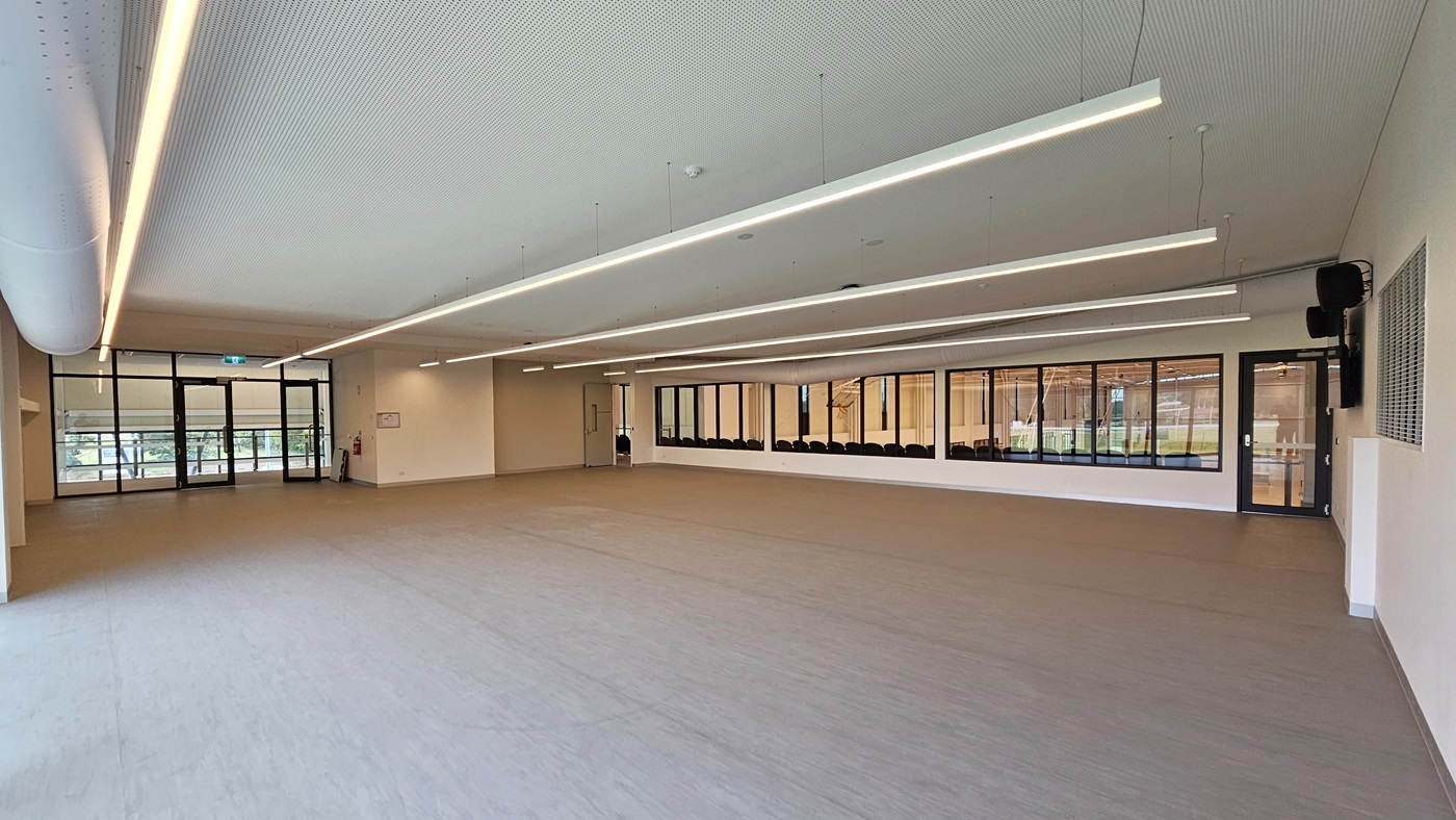 The Function Room is a spacious area with vinyl flooring. A large flat-screen TV and audio speakers are mounted on the right wall. Three doors provide access, two leading to the stadium's spectator area and another on the left leading to the lift. The walls are white, and the ceiling features a white porous design with fluorescent lighting and exposed large air conditioning vents. The Function Room is a spacious area with vinyl flooring. A large flat-screen TV and audio speakers are mounted on the right wall. Three doors provide access, two leading to the stadium's spectator area and another on the left leading to the lift. The walls are white, and the ceiling features a white porous design with fluorescent lighting and exposed large air conditioning vents.