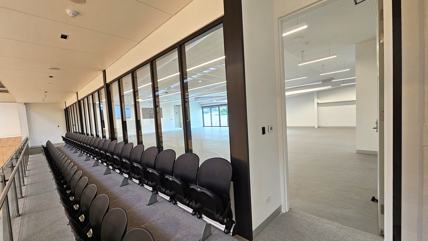 To the right is the Function Room, a spacious area with vinyl flooring. A large flat-screen TV and audio speakers are mounted on the right wall. Adjoining is the stadium's spectator area with chairs arranged for seating. A glass railing secures the area. The walls are white, and the ceiling features a white porous design with fluorescent lighting. To the right is the Function Room, a spacious area with vinyl flooring. A large flat-screen TV and audio speakers are mounted on the right wall. Adjoining is the stadium's spectator area with chairs arranged for seating. A glass railing secures the area. The walls are white, and the ceiling features a white porous design with fluorescent lighting.