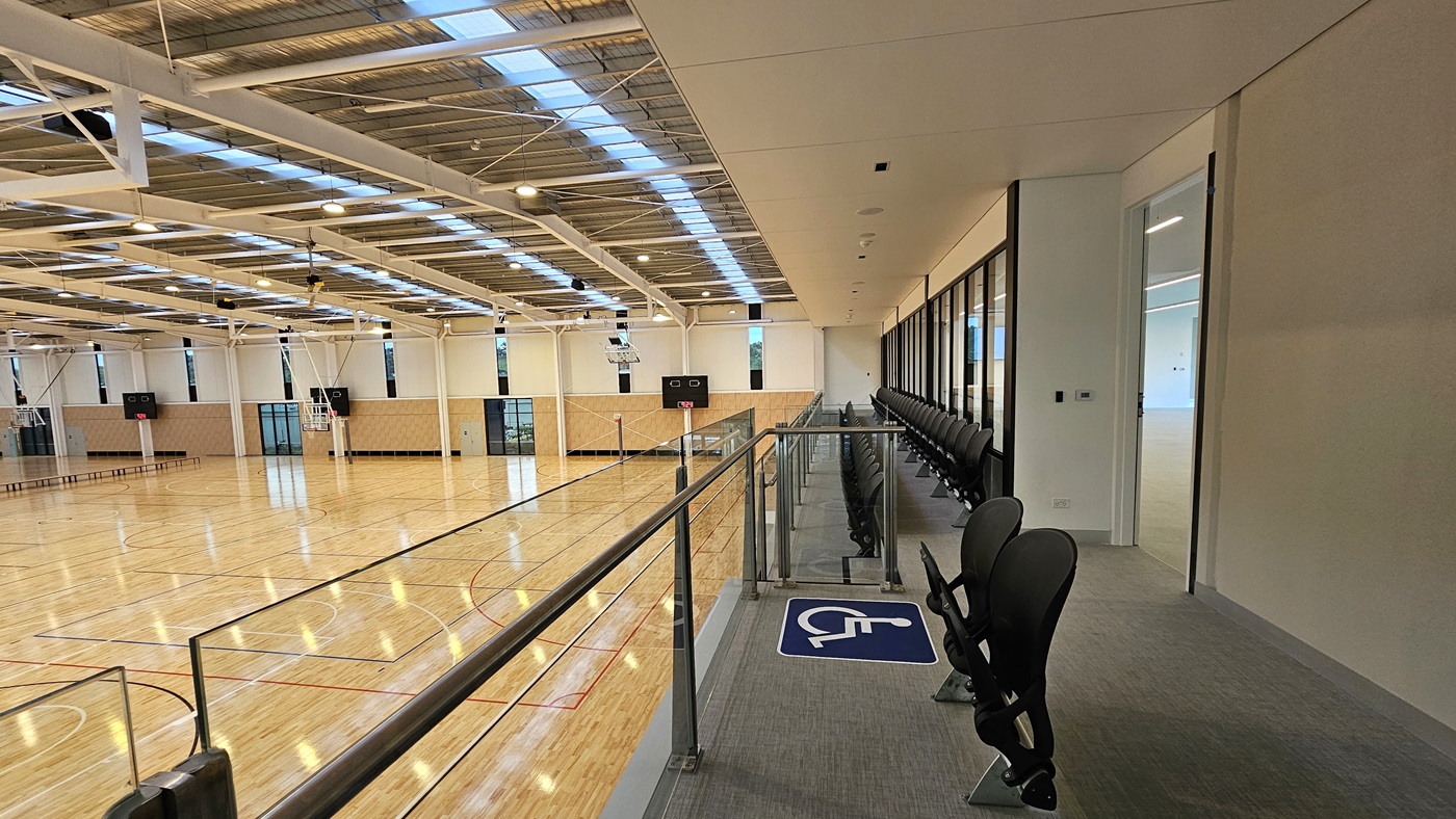 To the right is the Function Room, a spacious area with vinyl flooring. Adjoining is the stadium's spectator area with two rows of folded chairs for seating, including accessible seating. A glass railing secures the area. To the left is the stadium, featuring timber floors and an industrial ceiling with exposed beams, skylights, and aluminum insulation. To the right is the Function Room, a spacious area with vinyl flooring. Adjoining is the stadium's spectator area with two rows of folded chairs for seating, including accessible seating. A glass railing secures the area. To the left is the stadium, featuring timber floors and an industrial ceiling with exposed beams, skylights, and aluminum insulation.