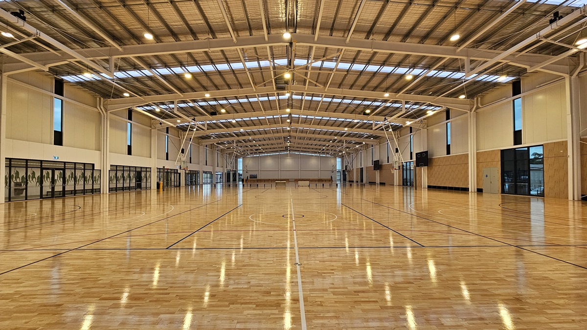The stadium is a spacious area with polished timber flooring marked for various ball games. The ceiling has an industrial aesthetic with exposed beams, aluminum insulation, and ample skylights. On the left, a mezzanine offers seating for spectators, surrounded by glass. The stadium is a spacious area with polished timber flooring marked for various ball games. The ceiling has an industrial aesthetic with exposed beams, aluminum insulation, and ample skylights. On the left, a mezzanine offers seating for spectators, surrounded by glass.