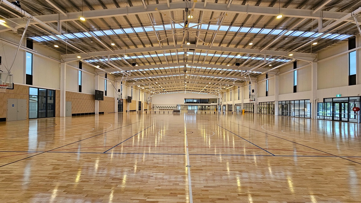 The stadium features a spacious area with polished timber flooring marked for basketball. Its industrial-style ceiling has exposed beams, aluminum insulation, and ample skylights. The wall behind is entirely made of glass. A fire hose reel and an "exit" sign are visible on the left. A basketball board is in the middle and on top of it is a digital screen. The stadium features a spacious area with polished timber flooring marked for basketball. Its industrial-style ceiling has exposed beams, aluminum insulation, and ample skylights. The wall behind is entirely made of glass. A fire hose reel and an "exit" sign are visible on the left. A basketball board is in the middle and on top of it is a digital screen.