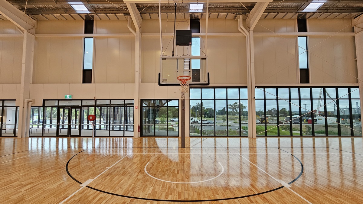 A spacious seating area overlooks the stadium with bench chairs positioned in the middle. The stadium, surrounded by glass, features timber flooring. At the far end is a glass door marked with an "Exit" sign. The floor is firm vinyl, and the ceiling has a white porous design with fluorescent lighting. A spacious seating area overlooks the stadium with bench chairs positioned in the middle. The stadium, surrounded by glass, features timber flooring. At the far end is a glass door marked with an "Exit" sign. The floor is firm vinyl, and the ceiling has a white porous design with fluorescent lighting.