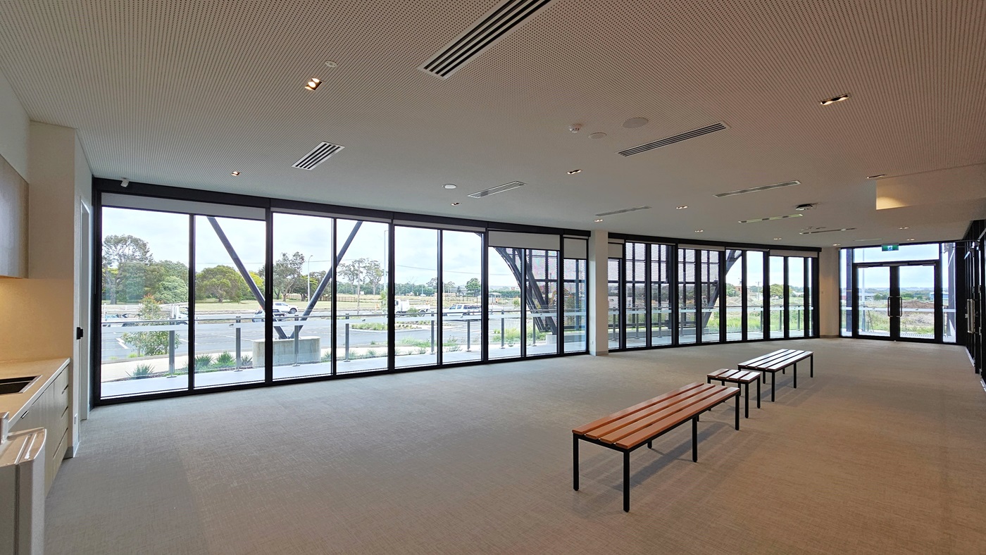 The mezzanine area features seats for spectators, including accessible seating, each marked. A stainless steel glass railing secures the area. From this view, the court below is visible, featuring polished timber floors and an industrial-look ceiling with exposed beams, aluminum insulation, and skylights. The mezzanine area features seats for spectators, including accessible seating, each marked. A stainless steel glass railing secures the area. From this view, the court below is visible, featuring polished timber floors and an industrial-look ceiling with exposed beams, aluminum insulation, and skylights.