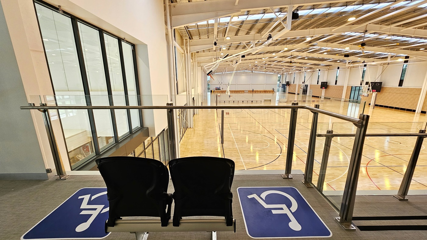 The mezzanine area has two rows of seats for spectators, including accessible seating, each marked. A stainless steel glass railing secures the area. From here, the court below is visible, with polished timber floors and an industrial-style ceiling featuring exposed beams, aluminum insulation, and skylights. The mezzanine area has two rows of seats for spectators, including accessible seating, each marked. A stainless steel glass railing secures the area. From here, the court below is visible, with polished timber floors and an industrial-style ceiling featuring exposed beams, aluminum insulation, and skylights.