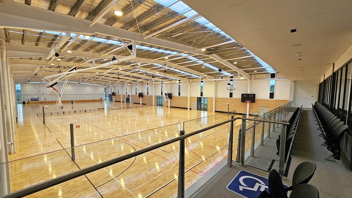 To the right is the stadium's spectator area with two rows of folded chairs for seating. A glass railing secures the area. To the left is the stadium, featuring timber floors marked for various ball games and an industrial ceiling with exposed beams, skylights, and aluminum insulation. To the right is the stadium's spectator area with two rows of folded chairs for seating. A glass railing secures the area. To the left is the stadium, featuring timber floors marked for various ball games and an industrial ceiling with exposed beams, skylights, and aluminum insulation.