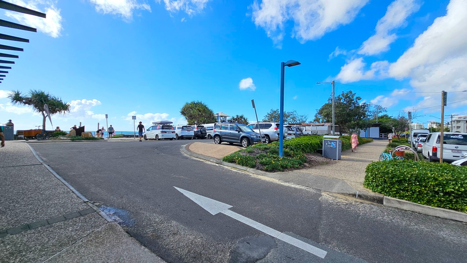 A paved area with a road in the centre featuring a white arrow pointing forward. On the left, a pedestrian path with a pergola structure and a tree. In the middle, several parked cars, including a white one with a roof rack. On the right, a footpath lined with green bushes and a person walking. A blue lamp post and a utility box are visible. In the background, more trees and a glimpse of the ocean.