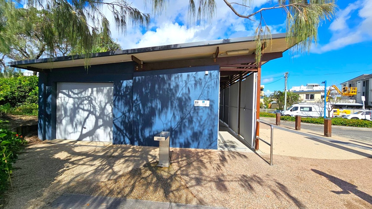 A public restroom building with a blue exterior wall and a white door on the left. Shadows of trees are cast on the wall and ground. A metal drinking fountain is in front of the wall. On the right, an open entrance with a metal railing leads inside. In the background, there are parked vehicles and buildings, including a construction site with a crane. The ground is paved with a textured surface, and trees with overhanging branches are visible.
