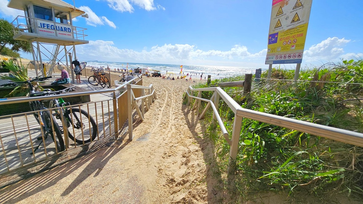 A sandy path leads to a beach with people and flags in the distance. On the left, a lifeguard tower stands on a wooden deck with bicycles parked nearby. A person sits on the deck, and another stands with a surfboard. On the right, a large sign with warnings and information is visible, surrounded by green vegetation. The ocean is in the background, with waves and a bright horizon.