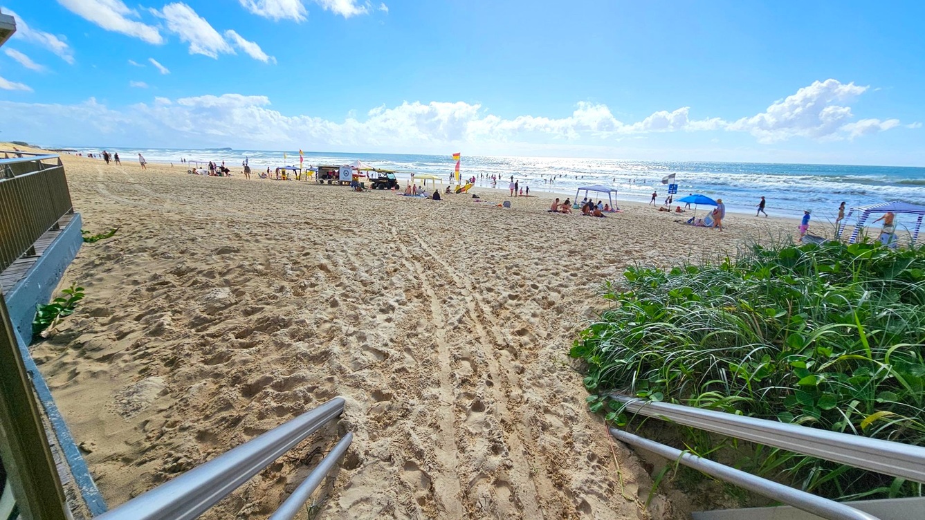 A sandy beach with people scattered across, some under umbrellas and tents. In the foreground, metal railings lead down to the sand. On the left, a building with a green railing is partially visible. On the right, there are green plants growing along the edge of the sand. The ocean is in the background, with waves approaching the shore and people swimming. The sky is blue with scattered clouds.