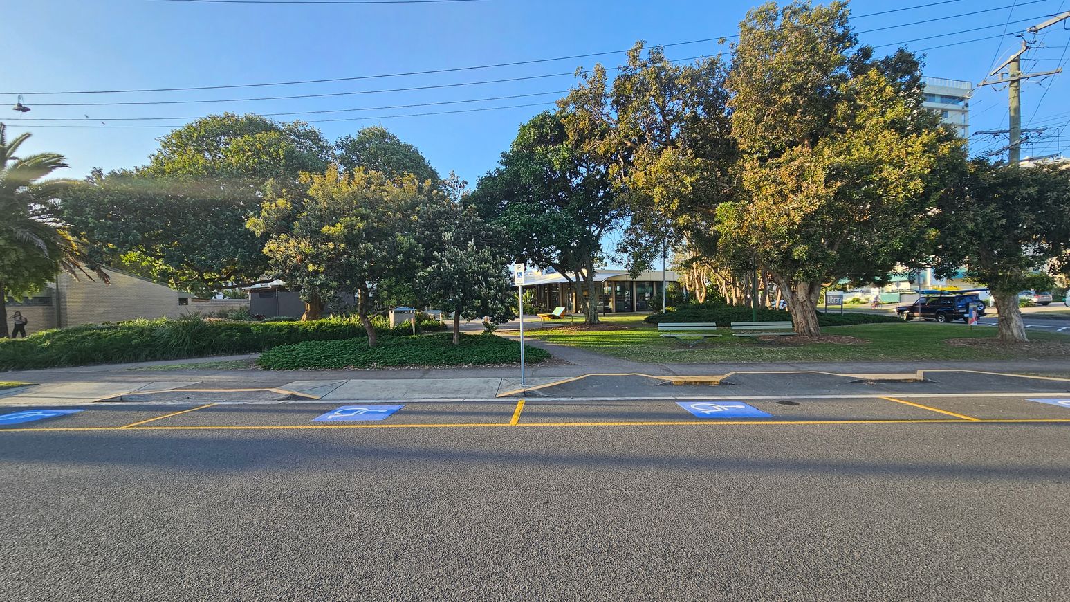 A street view with a row of trees and greenery in the background. A building with large windows is partially visible behind the trees. The foreground shows a road with marked parking spaces, including blue disabled parking signs. A pedestrian path runs parallel to the road, and a signpost is visible near the centre. A person is walking on the left side, and a vehicle is parked on the right. Overhead, power lines stretch across the scene.