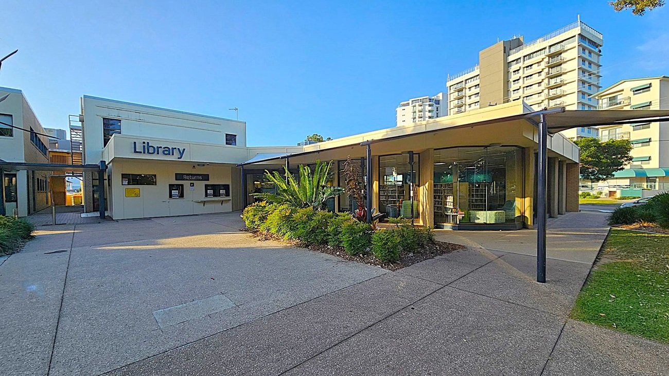 A modern library building with a white facade and large "Library" sign on the left. Below the sign are return slots. To the right, a curved glass section reveals bookshelves inside. In front, a landscaped area with green plants and a small tree. The ground is paved with concrete, and tall residential buildings are visible in the background. A metal canopy extends over the entrance path.