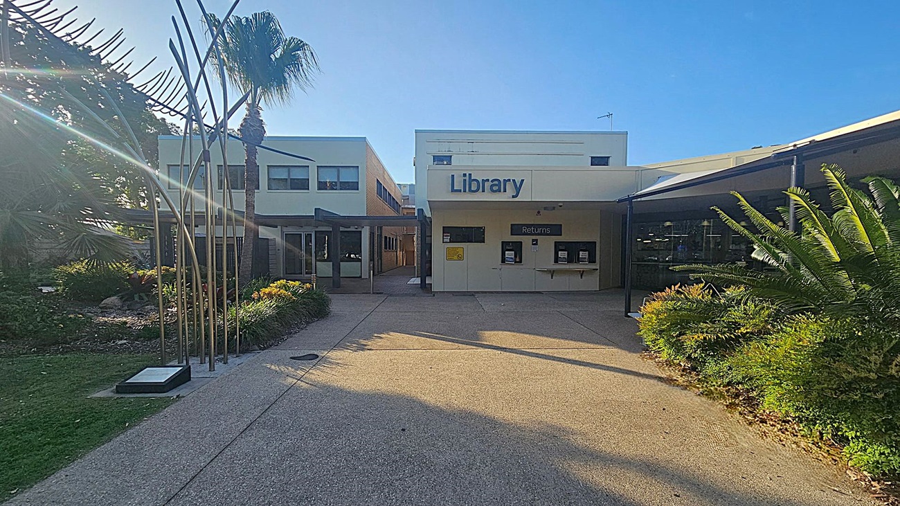 A library building with a sign reading "Library" above the entrance. To the left, there is a garden area with a palm tree and metal sculpture. The building has a white facade with a "Returns" section visible. On the right, there are more plants and a covered walkway. The ground is paved, and the area is well-lit by sunlight.