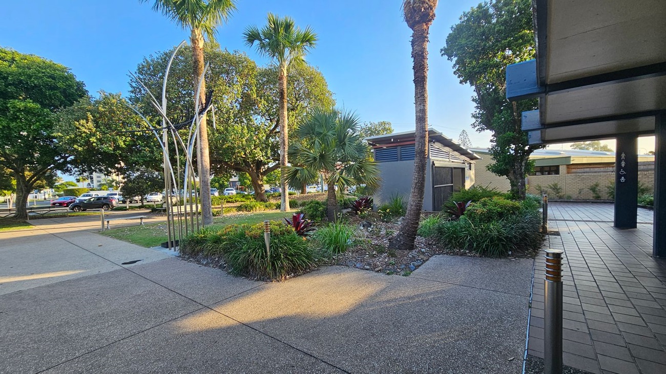 A paved walkway with a textured surface leads through a landscaped area featuring palm trees and various shrubs. On the left, a metal sculpture stands near a row of parked cars and large leafy trees. On the right, a building with a tiled entrance and a dark column is partially visible. The ground is a mix of concrete and brick paving, and the area is well-lit with small bollard lights.