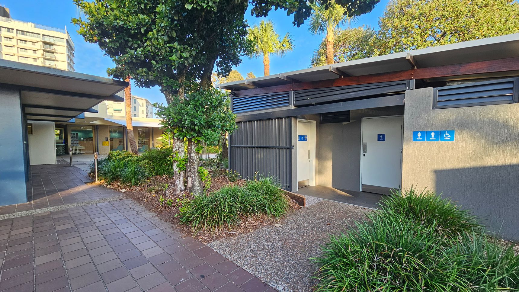 A public restroom building with a grey exterior is on the right, featuring signs for male, female, and accessible toilets. The entrance is surrounded by greenery, including bushes and a tree. On the left, there's a covered walkway with a tiled floor leading to another building. In the background, tall buildings are visible, and palm trees rise above the structures.