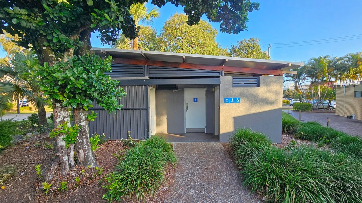 A public restroom building with a flat roof and grey walls is in the centre. The entrance has a door with a blue sign indicating accessibility. To the left, a large tree with dense green foliage stands next to the building. The foreground features a gravel path bordered by neatly trimmed grass and plants. In the background, there are more trees and a few parked cars. The area is well-lit, suggesting daytime.