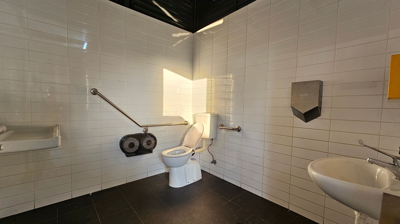 A public toilet with white tiled walls and a dark tiled floor. In the centre, there is a toilet with a white seat, equipped with metal grab bars on both sides. To the left, a sink with a soap dispenser is mounted on the wall. On the right, a hand dryer is installed above another sink. A large toilet paper dispenser is located near the toilet. The ceiling is dark, and sunlight casts a shadow on the wall.