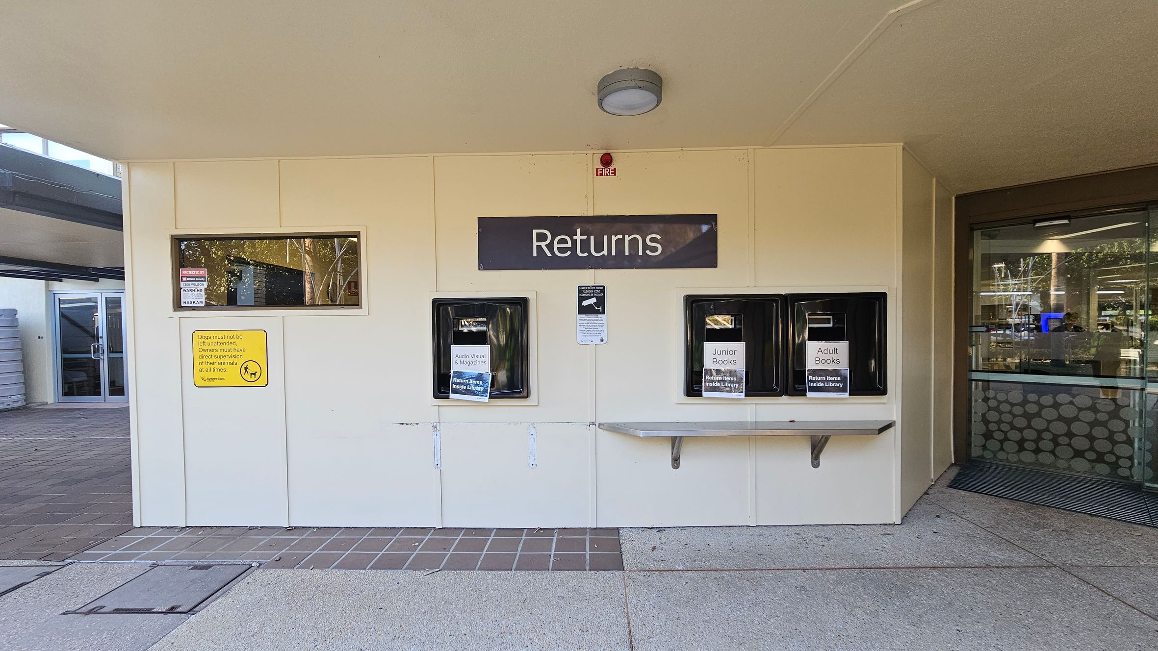 Entrance to a library with a glass door in the centre, displaying the name "Maroochydore Library". To the right, a glass panel lists opening hours and has several posters. Inside, bookshelves and a check-in desk are visible. The floor has a striped mat leading inside, and the ceiling is plain with a circular light fixture.