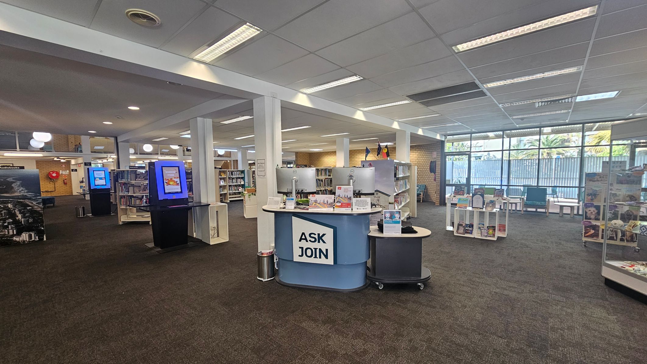 A library interior with a central information desk labelled "ASK JOIN" featuring two computer monitors. To the left, there are bookshelves and two digital kiosks with blue screens. The right side has a display shelf with books and a seating area near large windows. The floor is carpeted, and the ceiling has fluorescent lighting.
