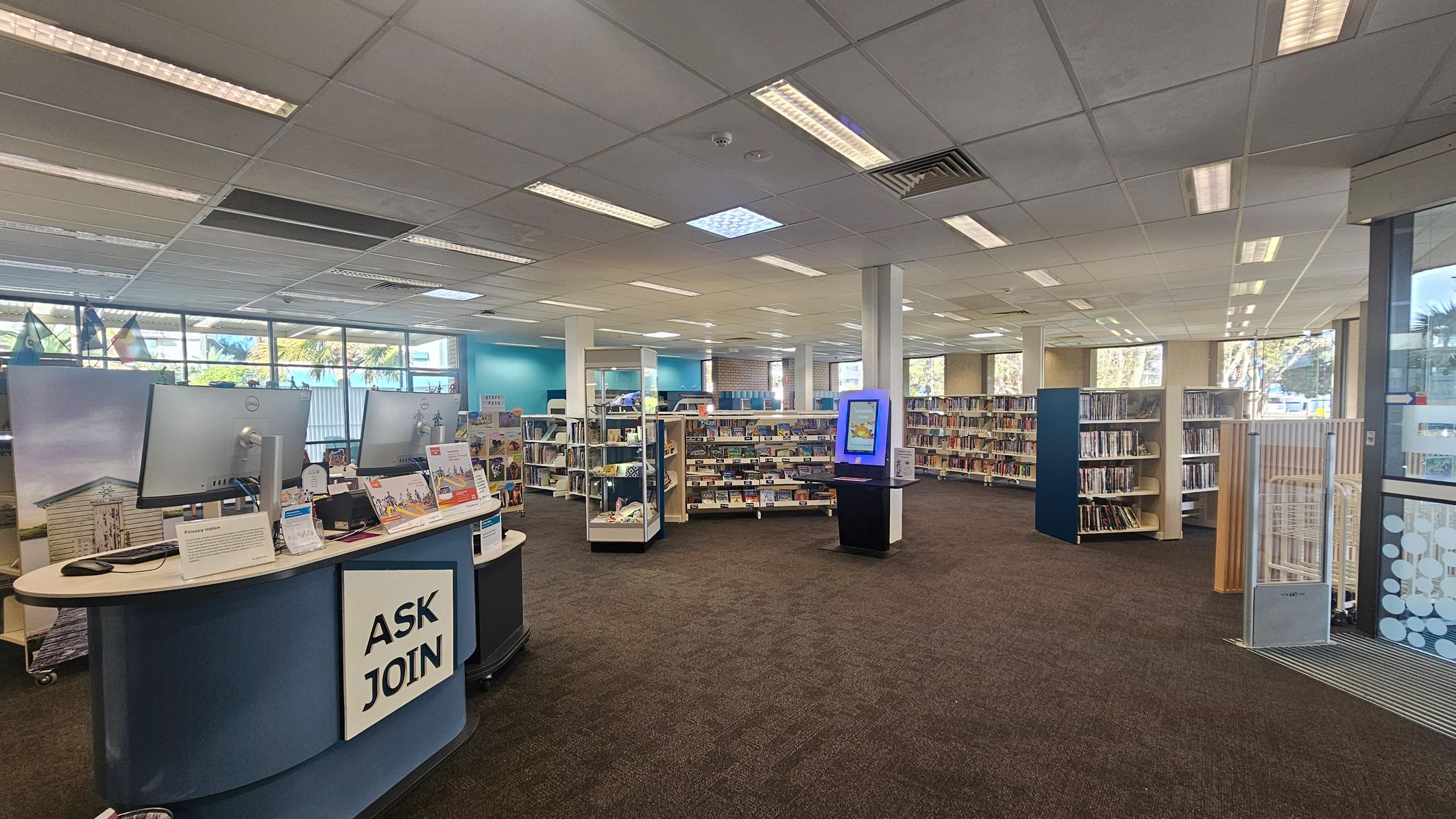 A library interior with a brown carpeted floor and a grid-patterned ceiling with fluorescent lights. On the left, there's a curved desk with two computer monitors and a sign reading "ASK JOIN." In the middle, several bookshelves are filled with books, and a digital kiosk stands nearby. The right side features more bookshelves and a glass entrance with a patterned design. Large windows in the background allow natural light to enter, with some greenery visible outside.