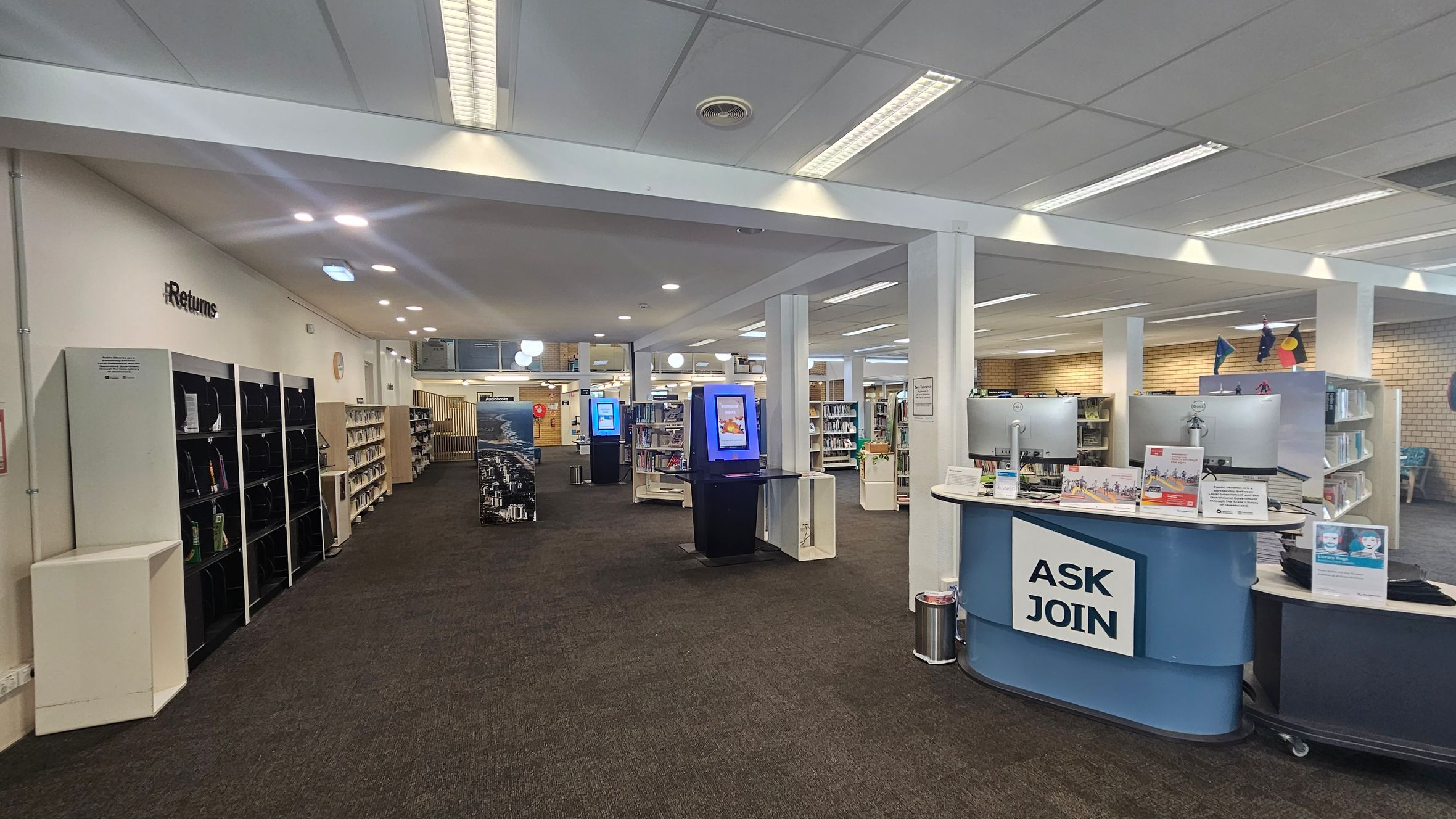 Library interior with a brown carpeted floor. On the left, there are shelves labelled "Returns" with books. In the centre, a blue kiosk with a screen stands among more bookshelves. On the right, a curved desk with "ASK JOIN" signage, two computer monitors, and informational pamphlets. The ceiling is white with fluorescent lights. In the background, more bookshelves and a wall with flags.