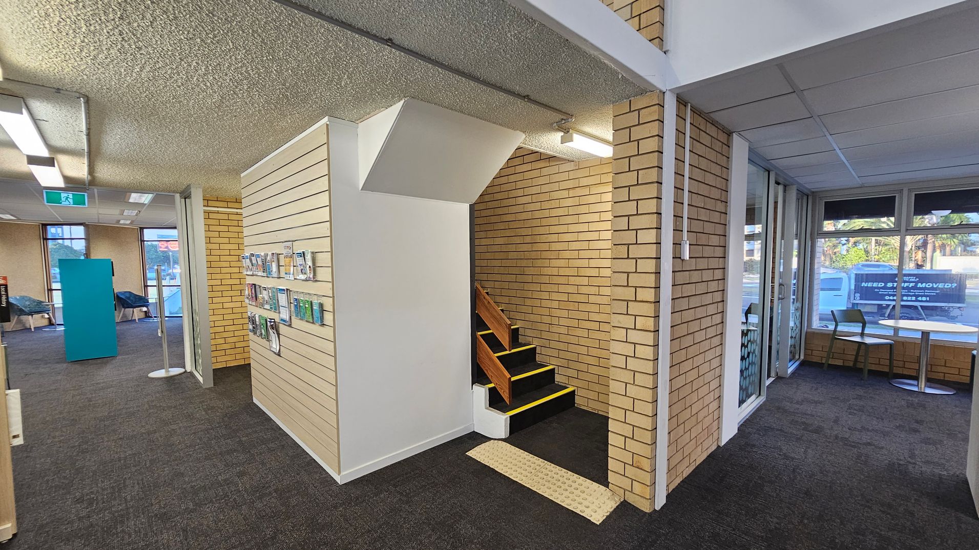 On the left, there is a desk with a computer and two green chairs. Adjacent to it, a wooden slatted partition separates the area. In the middle, two blue chairs are placed against the wall beneath framed pictures. On the right, an elevator door is visible next to a brick wall with a red fire hose reel and a fire extinguisher mounted on it. The floor is carpeted in dark grey, and the ceiling has fluorescent lights.