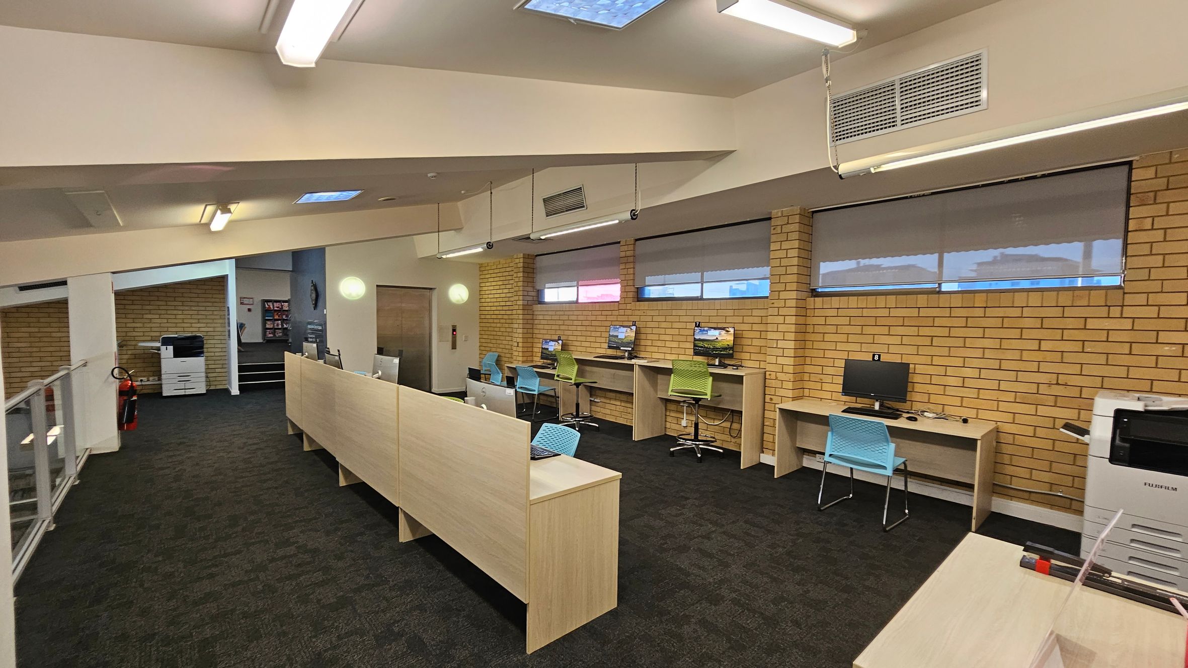 An office space with a row of desks and computers along a brick wall on the right. Each desk has a chair, some blue and some green. On the left, there is a glass railing and a large printer near a fire extinguisher. The floor is carpeted in dark tones, and the ceiling has fluorescent lights and a skylight. The walls are a mix of brick and white surfaces.