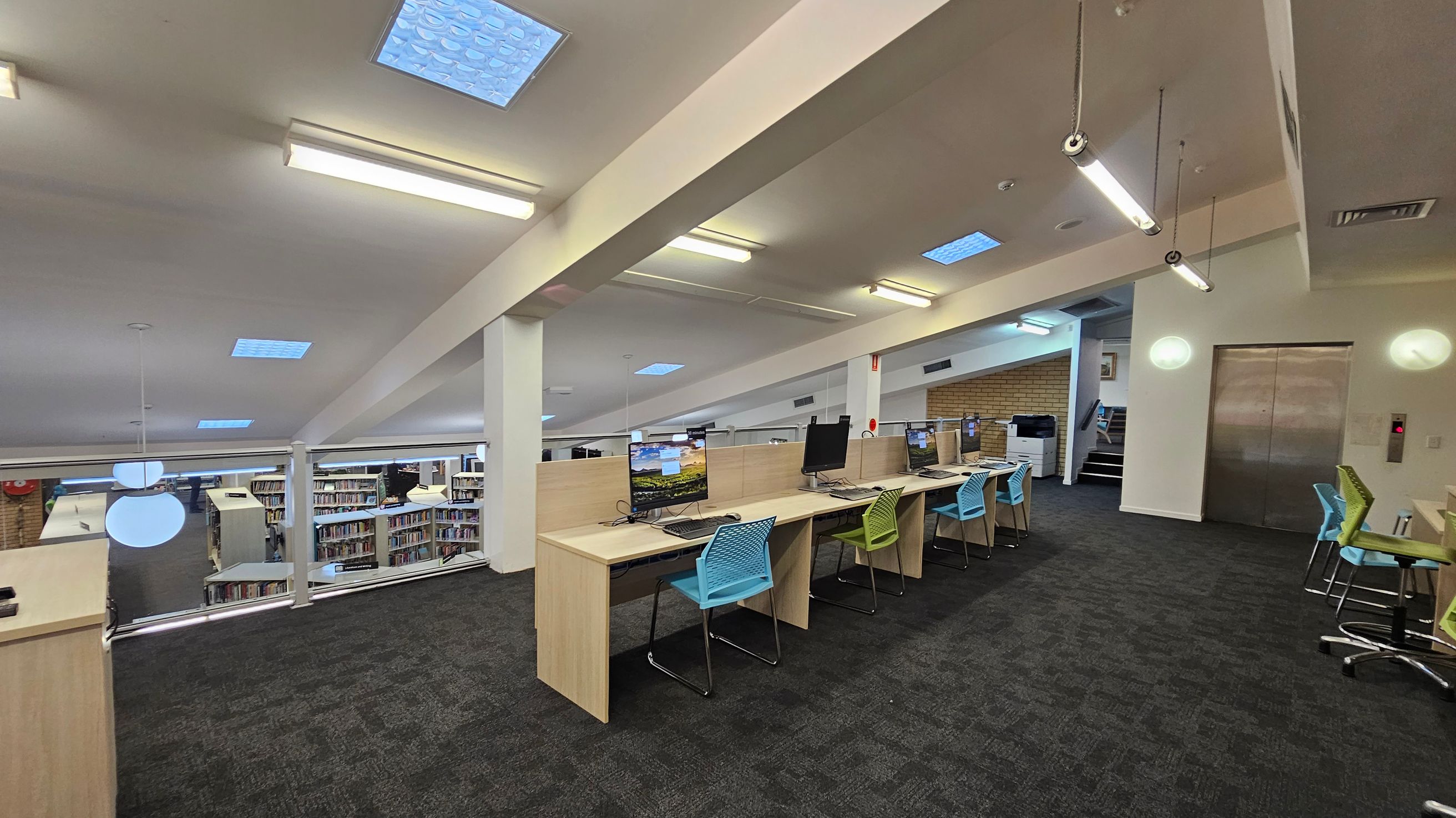 A modern library interior with a row of computer desks on the right, each equipped with a monitor and colourful chairs. The floor is carpeted in dark grey. On the left, there is a glass barrier overlooking bookshelves filled with books. The ceiling features fluorescent lights and skylights. In the background, there is a lift with metallic doors and a small staircase leading to another level.