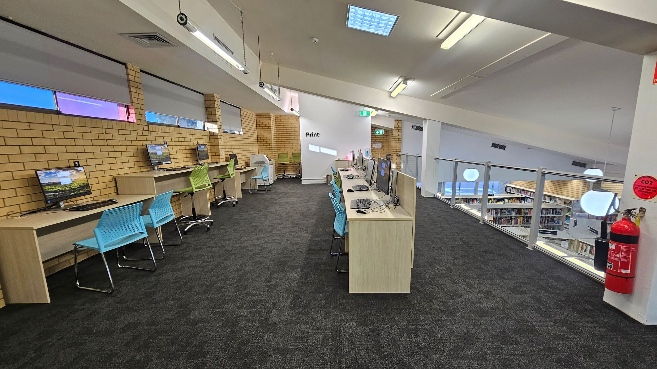 A library interior with a row of computers on wooden desks along the left wall, accompanied by blue and green chairs. The wall is made of light brown bricks with small windows above. In the centre, more computers are on a long desk with blue chairs. A white wall with "Print" written on it is at the back. On the right, a glass railing overlooks bookshelves below. A fire extinguisher is mounted on the wall. The floor is carpeted in dark grey, and the ceiling has fluorescent lights.