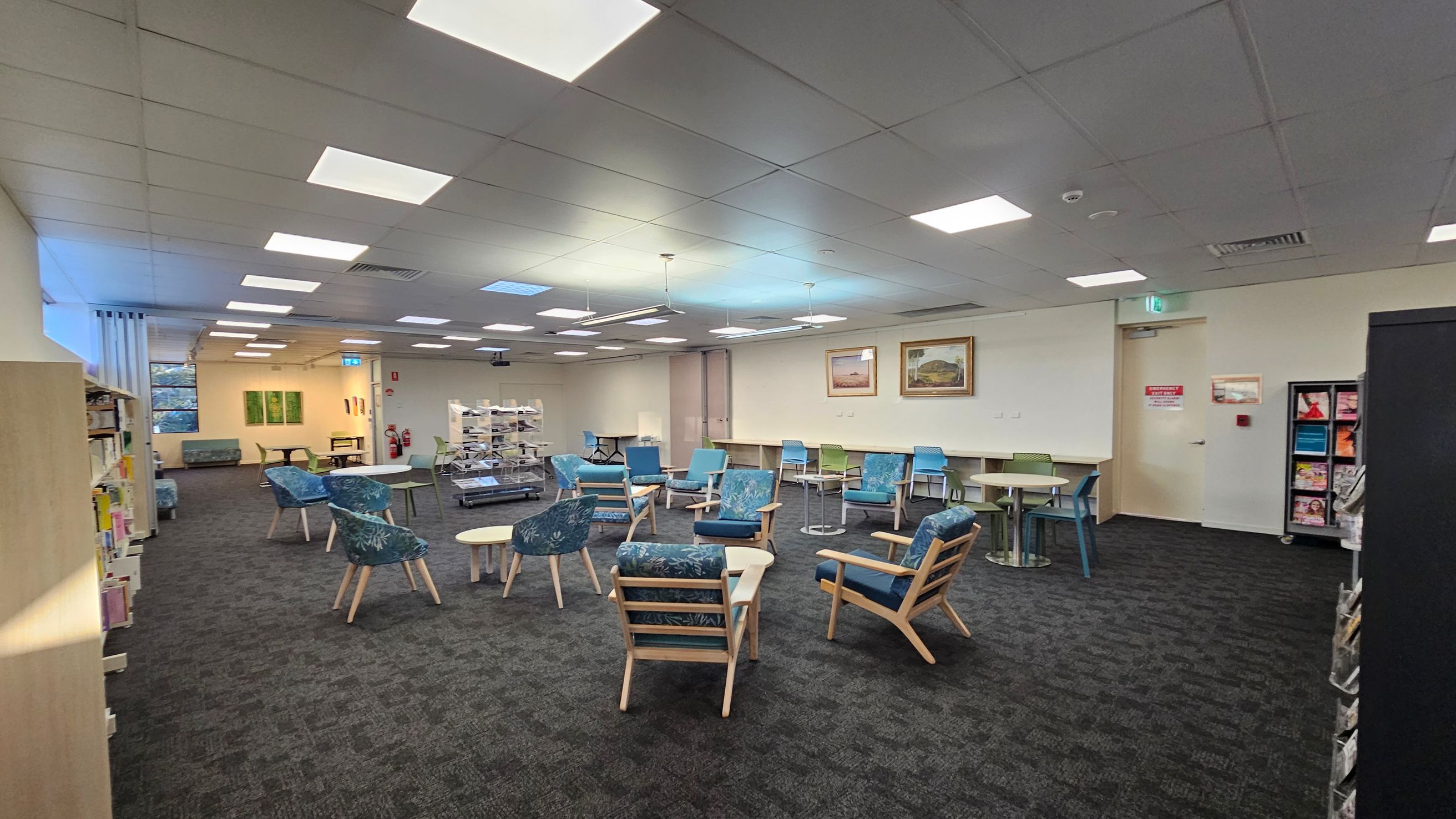 A spacious room with a dark carpeted floor and a grid-patterned ceiling with fluorescent lights. In the centre, there are several blue and green cushioned chairs and small round tables. On the left, bookshelves line the wall. The right side features a magazine rack and a door with a red sign. The back wall has framed paintings and a row of tables with chairs. A fire extinguisher is visible near the back. The room has a calm, organised layout.