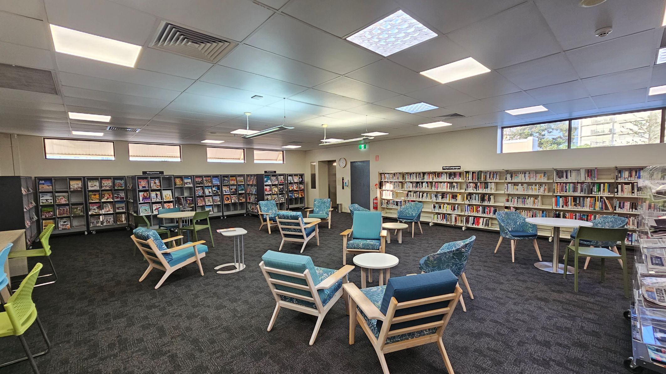 A library interior with a dark carpeted floor and a ceiling with recessed lighting. In the centre, there are several wooden chairs with blue cushions and small round tables. On the left, there are shelves filled with magazines and a few green chairs at tables. On the right, bookshelves are lined with books, labelled "World Languages." Large windows near the ceiling allow natural light in. A clock and a fire extinguisher are visible on the back wall.