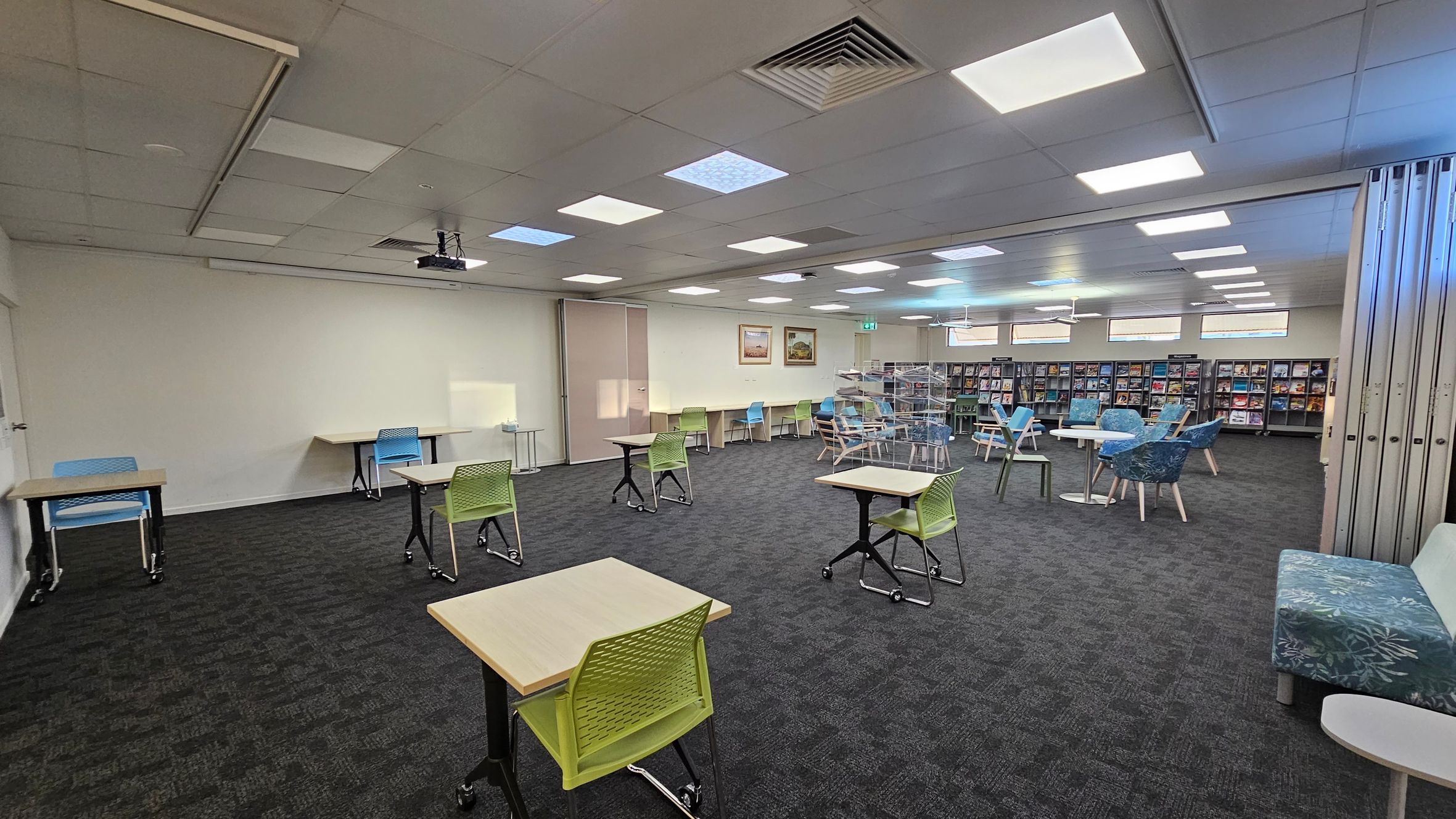 A spacious room with a dark carpeted floor and a ceiling with recessed lighting. On the left, there are individual desks with green and blue chairs. In the middle, more desks and chairs are scattered, with a projector mounted on the ceiling. On the right, there are bookshelves filled with books and magazines, along with round tables and blue patterned chairs. A floral-patterned bench is also visible. The walls are light-coloured, and there are a few framed pictures.