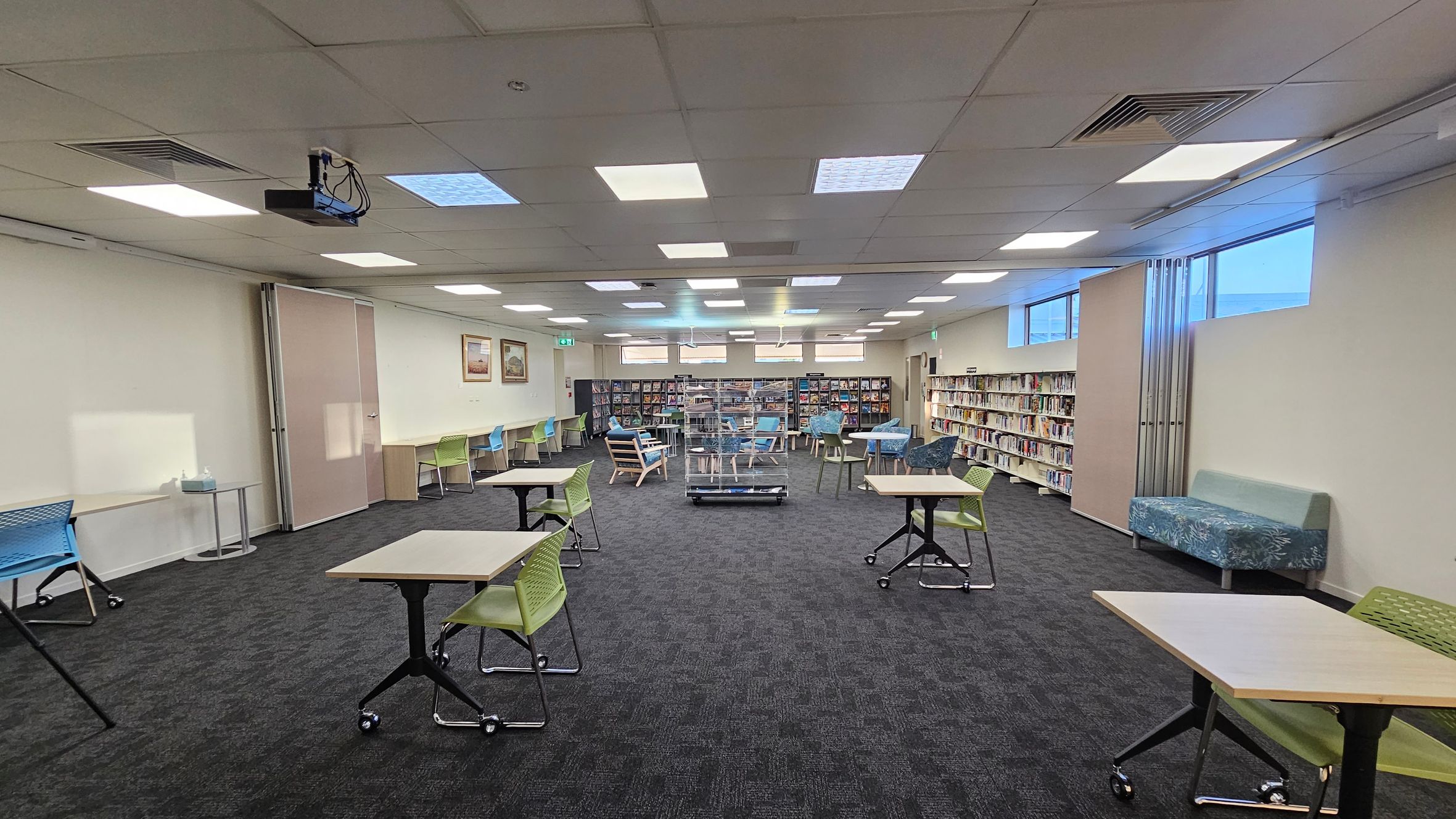A spacious library room with a dark carpeted floor and a ceiling with recessed lighting. In the foreground, there are tables with green chairs on wheels. The left side has a small round table and a blue chair. The right side features a patterned sofa and bookshelves filled with books. The back wall has more bookshelves and a few framed pictures. There are windows high on the walls, and a projector is mounted on the ceiling.