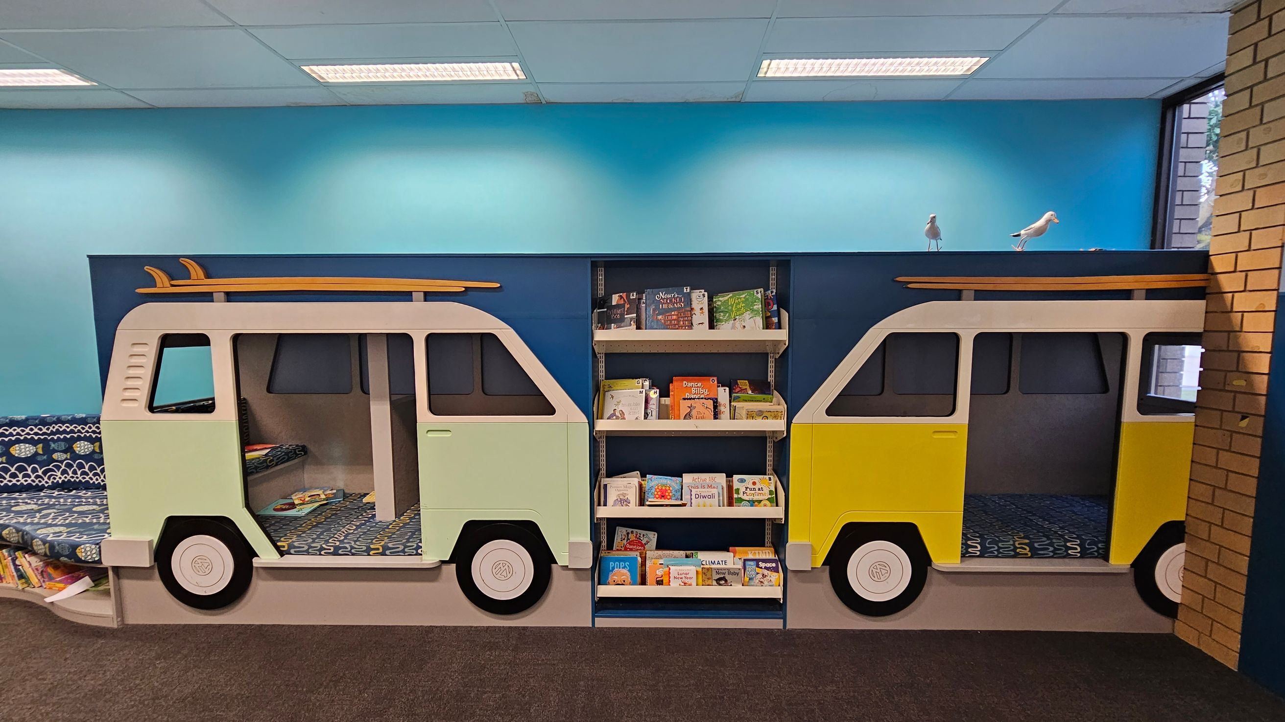 A children's reading area with a playful design featuring two van-shaped reading nooks. The left van is light green, and the right van is yellow, both with surfboards on top. Between them is a bookshelf filled with children's books. The background wall is painted blue, and there are two bird figurines on top of the structure. The floor is carpeted, and there's a brick wall on the right side.