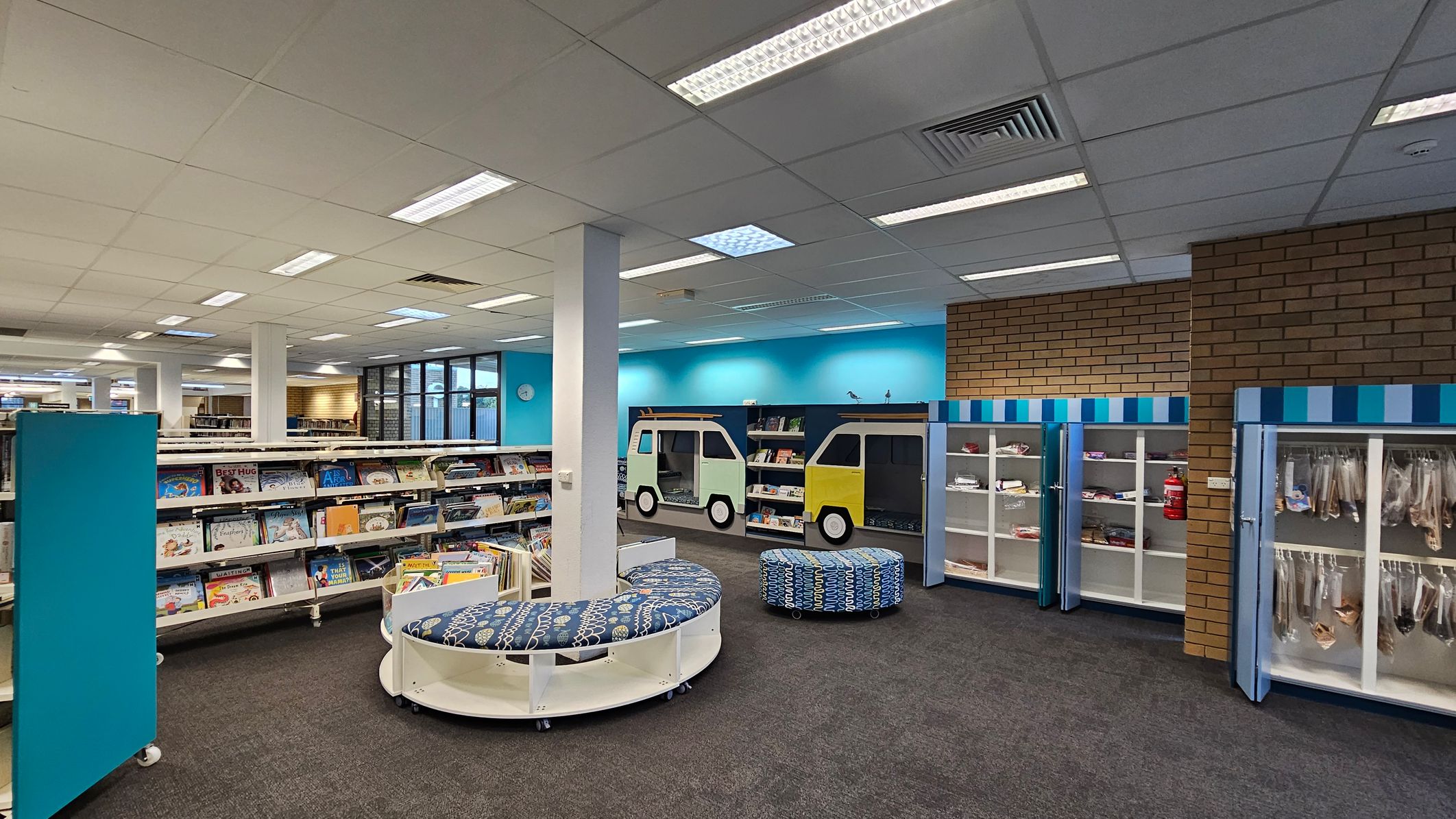 A library interior with a grey carpeted floor and a white tiled ceiling with fluorescent lights. On the left, there are shelves filled with children's books. In the centre, there are circular, patterned seating areas. The right side features a brick wall and display cabinets with blue and white striped tops. The back wall is painted blue with windows and a clock. There are also bookcases designed to look like vehicles.