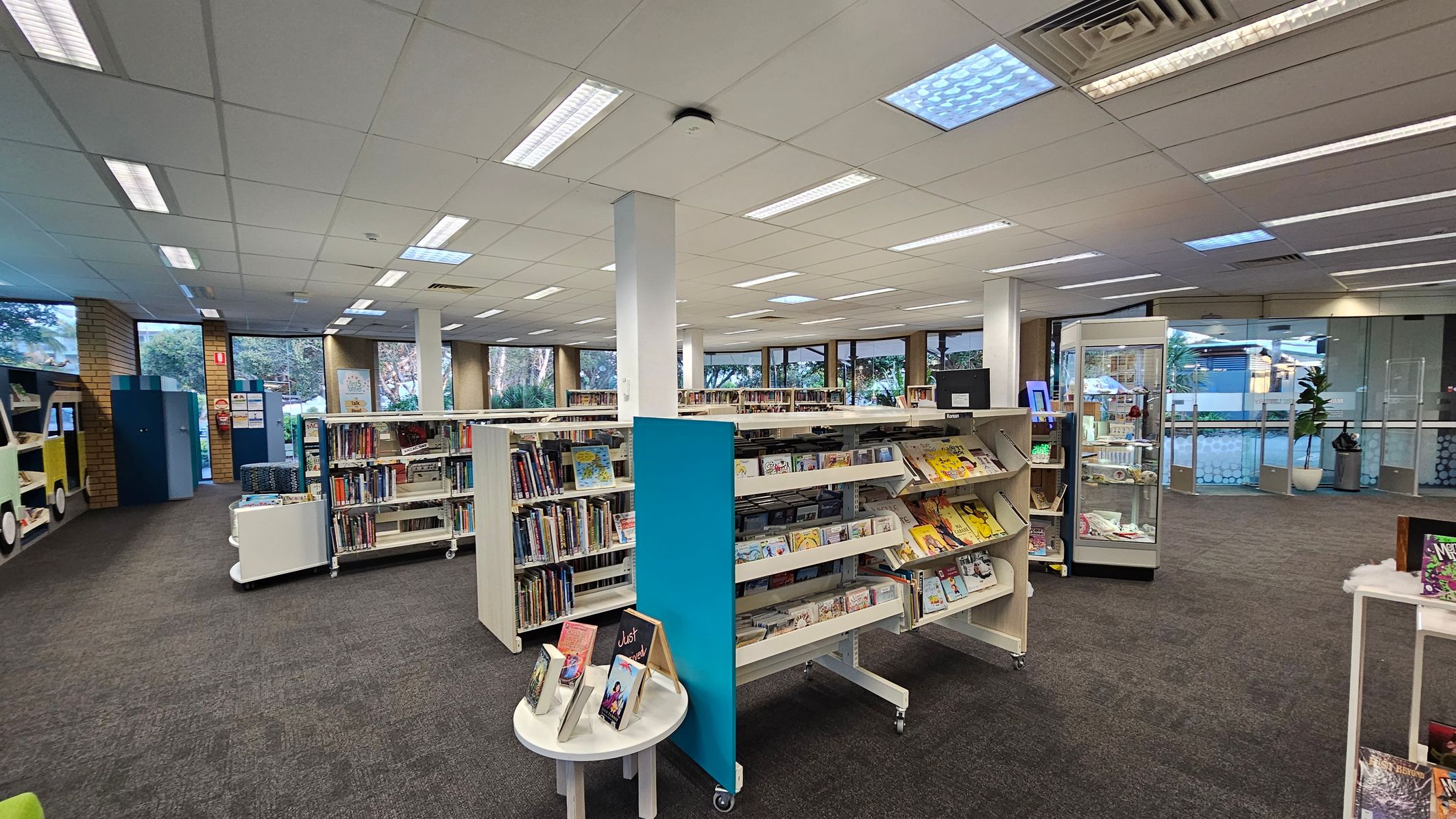 A library interior with grey carpeted flooring and a white panelled ceiling with fluorescent lights. In the centre, there are several white bookshelves filled with books and magazines. On the left, a colourful book display shaped like a vehicle. On the right, a glass display case and a potted plant near the entrance. Large windows in the background provide natural light and a view of greenery outside.