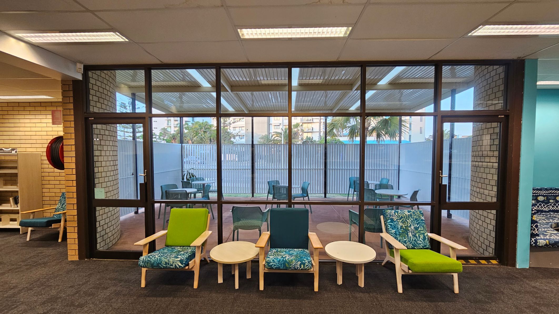 A room with a glass wall and door leading to an outdoor area. Inside, there are three wooden chairs with colourful cushions and two small round tables. The floor is carpeted in dark grey. Outside, there are several blue chairs and tables on a patio, with a view of palm trees and buildings in the background. The ceiling has fluorescent lights and the walls are made of light-coloured bricks.