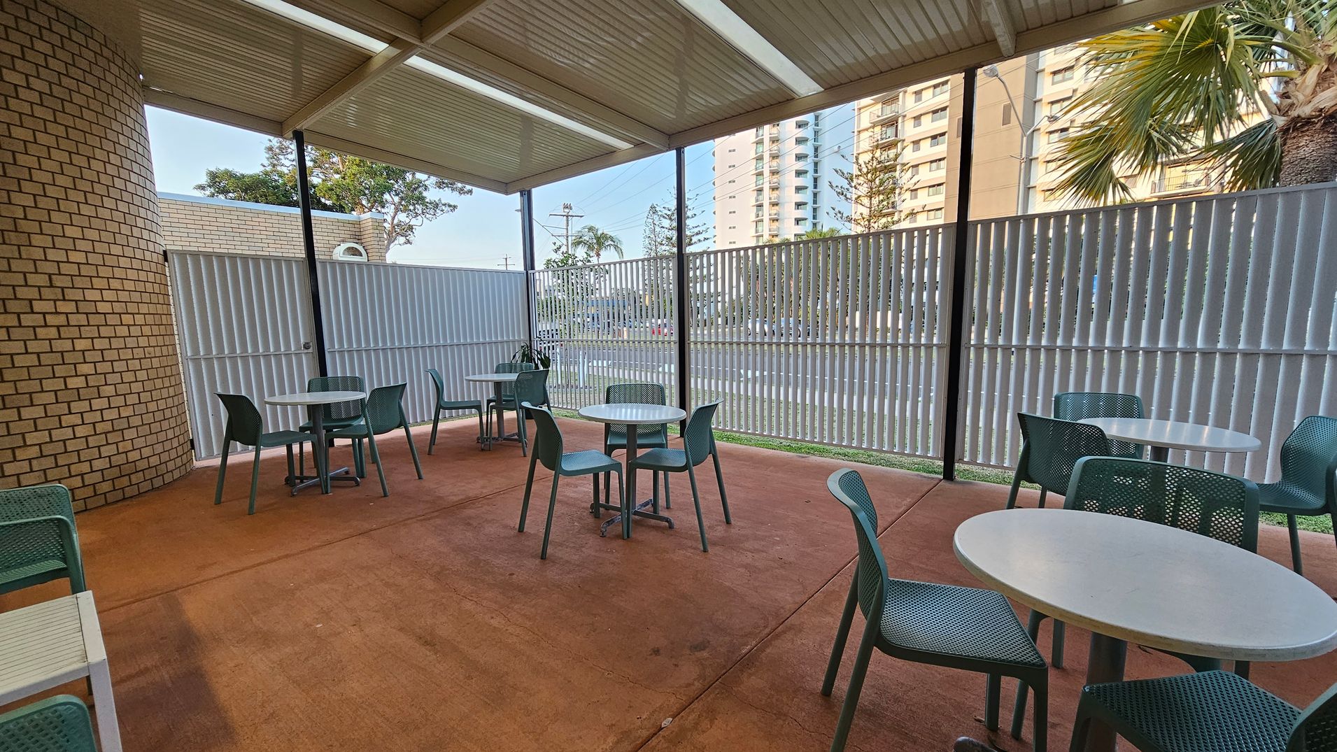 Outdoor seating area with several round tables and green chairs on a reddish-brown concrete floor. The left side features a curved brick wall, while the right side has a metal fence with vertical slats. In the background, there are tall buildings and palm trees visible through the fence. The ceiling is made of corrugated metal panels.