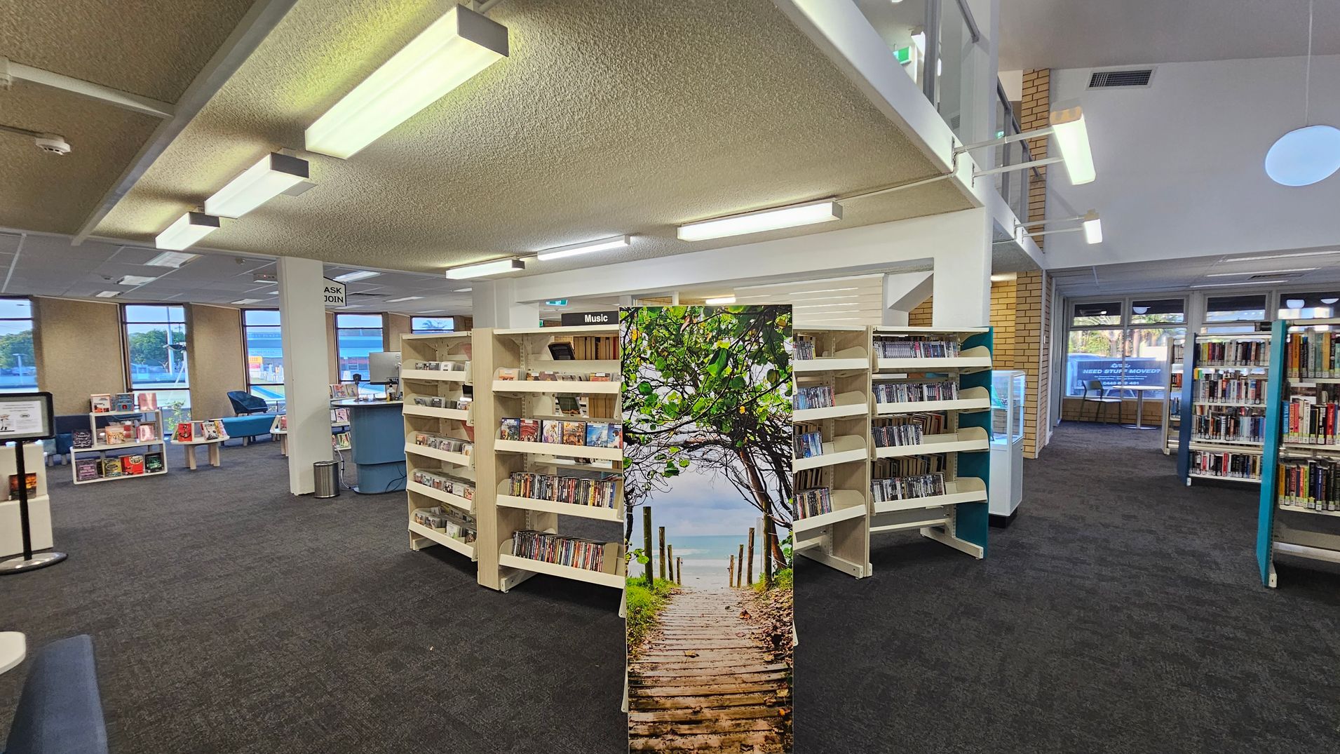 A library interior with a dark carpeted floor and a textured ceiling with fluorescent lights. In the centre, there are bookshelves filled with DVDs and CDs, labelled "Music". A decorative panel with an image of a wooden path leading to the sea is in front of the shelves. To the left, there are windows, a seating area with blue chairs, and a small desk. On the right, more bookshelves are visible, and large windows in the background let in natural light.